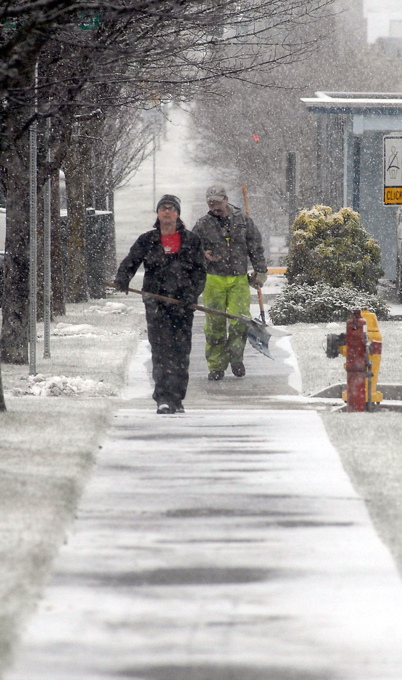 Port Angeles Parks Department workers Eli Hammel, left, and Leon Leonard carry snow shovels along a snowy stretch of sidewalk on East Fifth Street in front of Port Angeles City Hall on Tuesday. (Keith Thorpe/Peninsula Daily News)