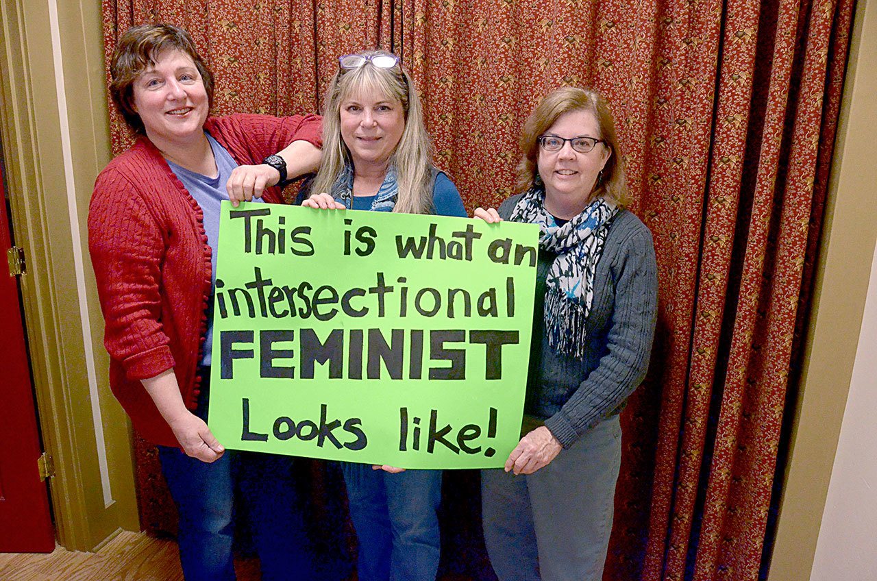 Robin Stemen, Susan Sherman and Annemarie Mende hold up one of the signs they’re taking to the Womxn’s March in Seattle on Saturday. (Cydney McFarland/Peninsula Daily News)