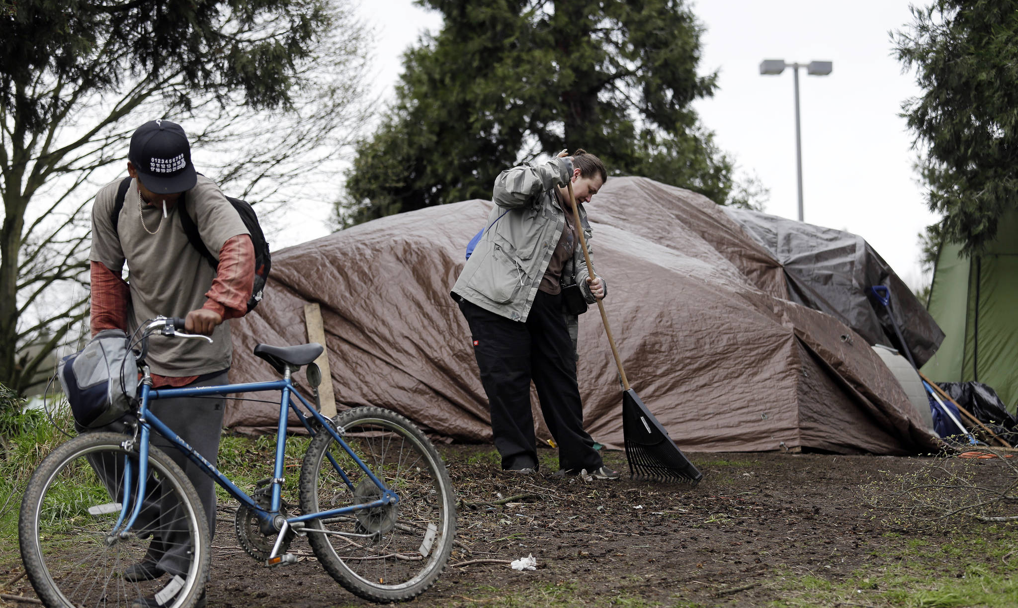 In this photo taken March 23, Lisa Hooper rakes debris from in front of the tent where she lives in a greenbelt near a freeway, as another camper wheels a bicycle past her in Seattle. (Elaine Thompson/The Associated Press)