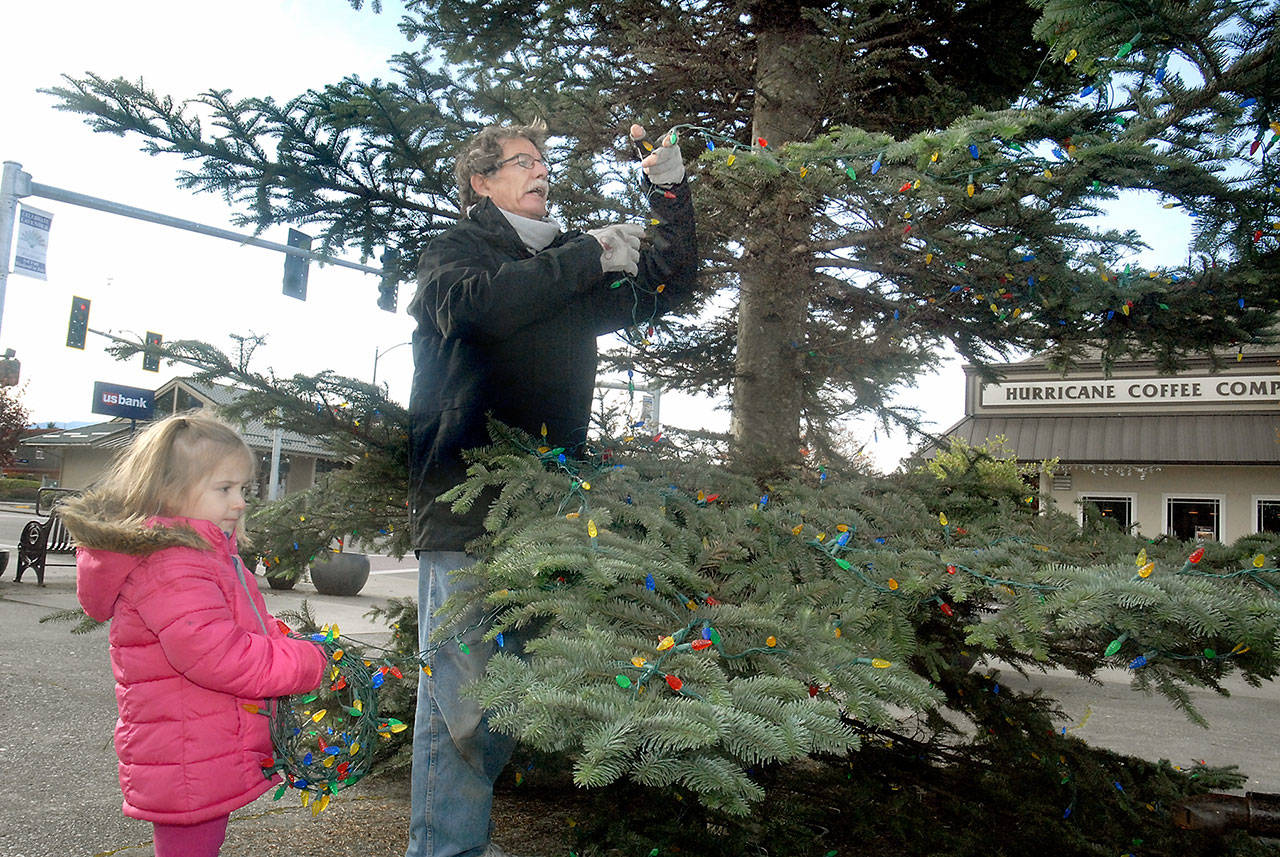 Rose Garcia, 4, of Port Angeles holds a string of Christmas lights while assisting Jeff Perry, a crew chief for Sequim-based Morning Star Balloon Co., as he decorates the city Christmas tree at Washington Street and Sequim Avenue in Sequim on Saturday. The tree will be the centerpiece when Santa Claus arrives at the location next Saturday for a community holiday celebration. (Keith Thorpe/Peninsula Daily News)