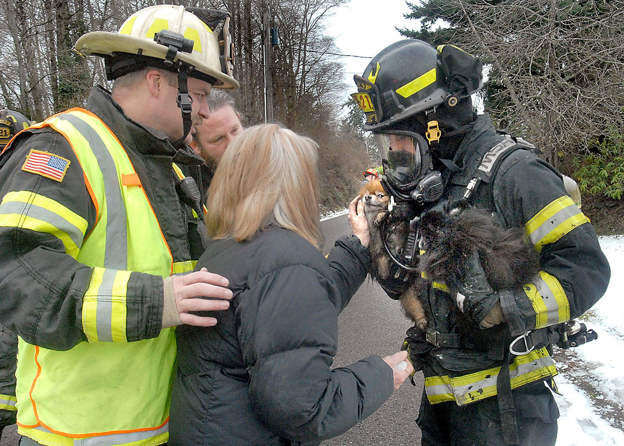 Fire victim Judy Zeeb, center, is reunited with two of her dogs held by Clallam County Fire District No. 2 firefighter Michael Stroobant, right, as Deputy Chief Jake Patterson, left, looks on while other firefighters extinguish a blaze in Zeeb’s East Bay Street home east of Port Angeles on Wednesday. (Keith Thorpe/Peninsula Daily News)