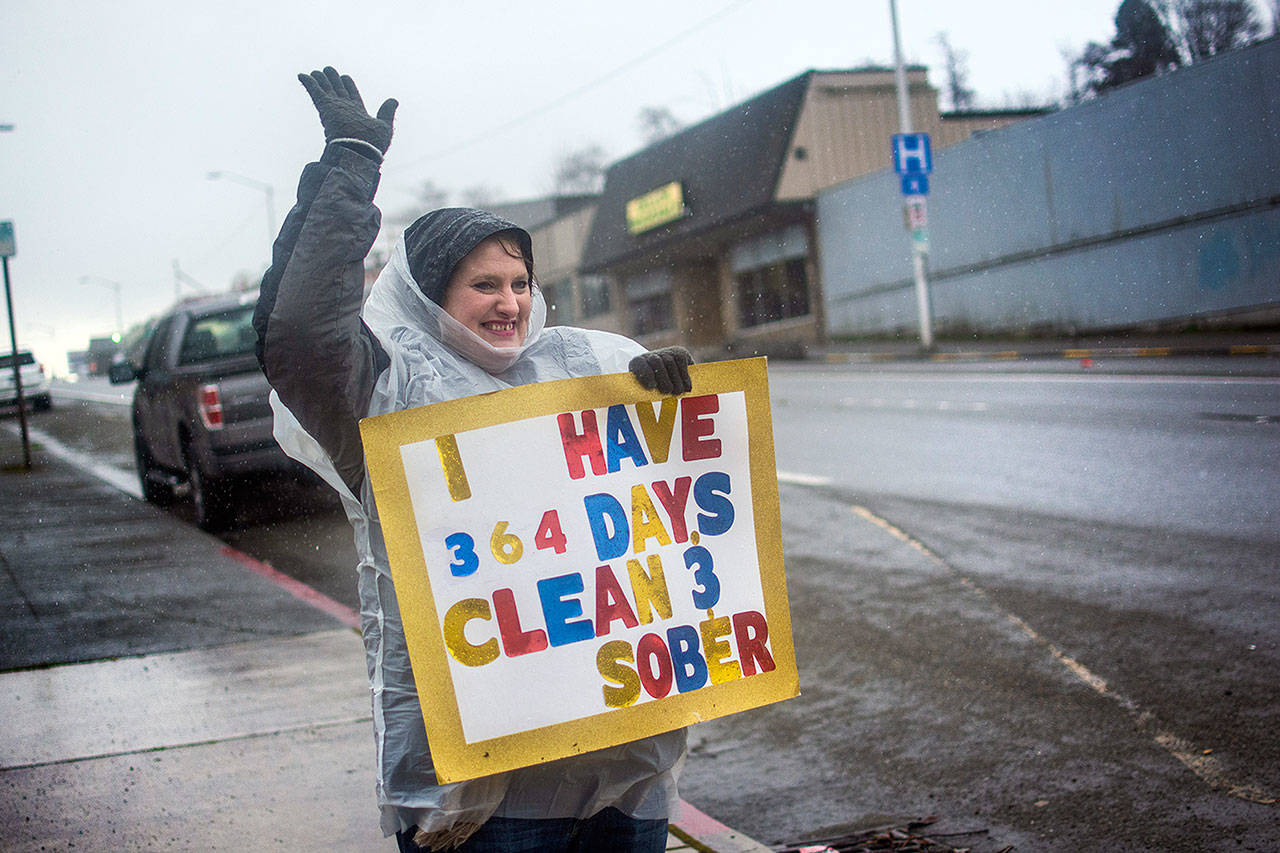 Jenni Tiderman stands in the pouring rain at the corner of First and Lincoln Streets in Port Angeles on Friday with a sign declaring she is 364 days sober. Over the last year she has secured housing and a job. She also is regaining custody of her children. (Jesse Major/Peninsula Daily News)