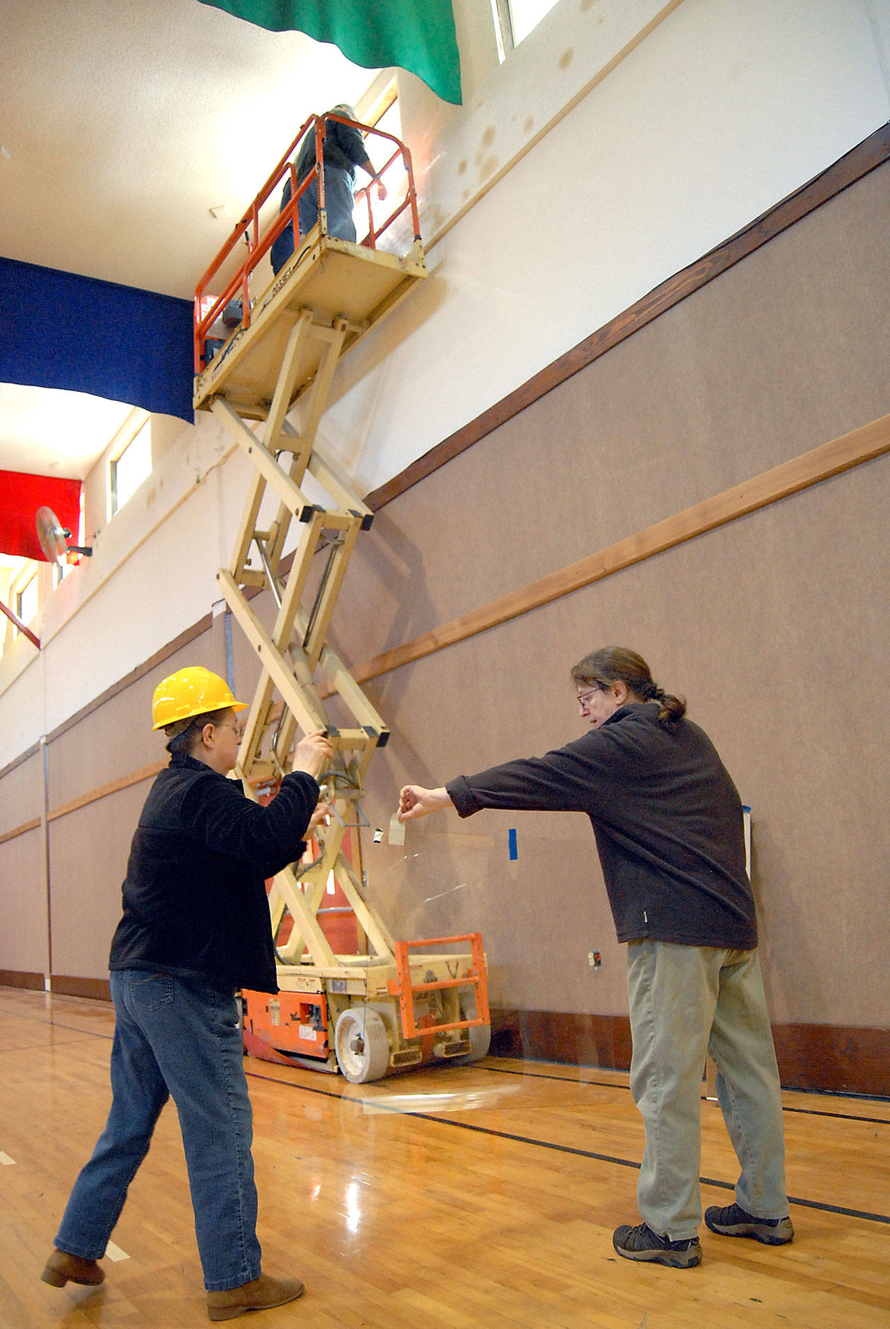 Marylan Thayer, left, and Rae Leiper prepare a piece of nearly invisible plastic window coating for installation in the gymnasium at the Joyce Bible Church in Joyce to help mitigate damage that could be caused by an earthquake. John Kent and Bruce Leiper, on lift, perform the installation. (Keith Thorpe/Peninsula Daily News)