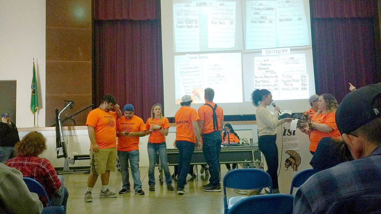 Anastasia Rigby announces information about an item being introduced for auction. To her left are four Spartan senior boys who volunteered 22 hours of work together to be auctioned off. On the screen in the background is a projection of bids in process. (Zorina Barker/for Peninsula Daily News)
