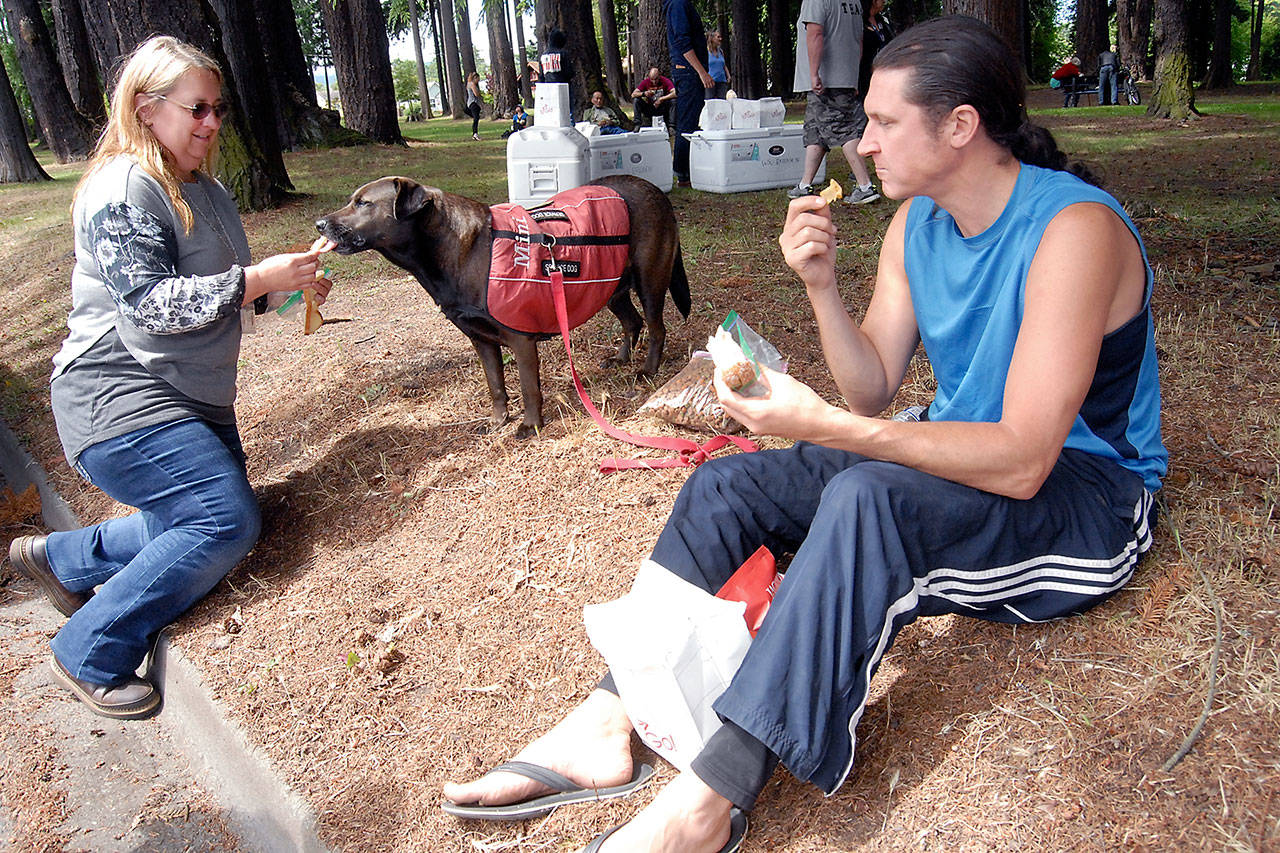 Rod Alger eats lunch as he watches Jaymie Doane, a case manager with Peninsula Behavioral Health, left, provide a snack for his dog, Mini, at Jesse Webster Park in Port Angeles. (Keith Thorpe/Peninsula Daily News)