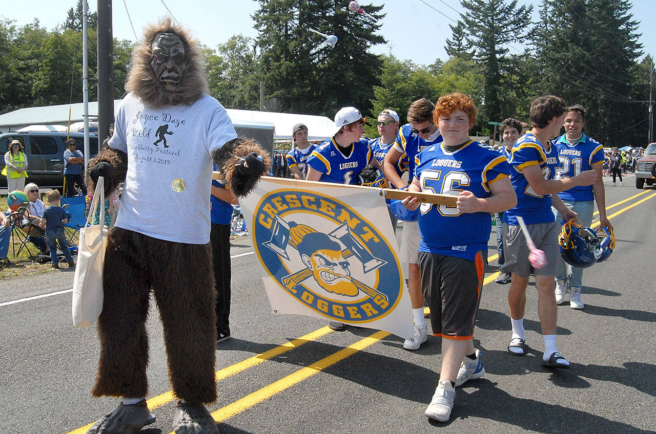 A sasquatch character, the mascot of the 2019 Joyce Daze Wild Blackberry Festival, tosses out candy to parade-goers while accompanied by members of the Crescent Loggers football team.