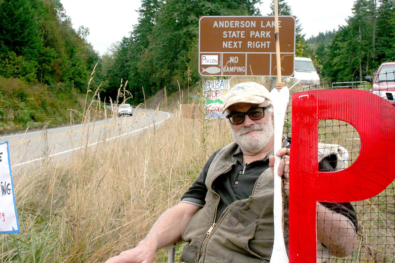 Norm Norton of Eaglemount sits in a chair in front of the Anderson Lake State Park sign along state Highway 20 on Monday. The protest group from the Jefferson County Environmental Coalition added a sign that reads “poison” above the fish on the state park sign. (Brian McLean/Peninsula Daily News)