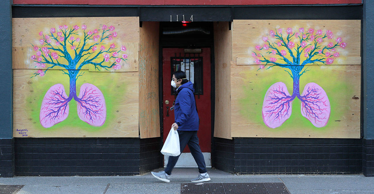 A pedestrian wearing a mask walks past paintings featuring lungs and a tree by Burgandy Viscosi on the boarded-up Re-Bar nightclub and theatre Tuesday, April 14, 2020, in downtown Seattle. Streets and sidewalks in the area were quiet Tuesday as most people in the city are staying and working at home due to the outbreak of the coronavirus. (Ted S. Warren/The Associated Press)
