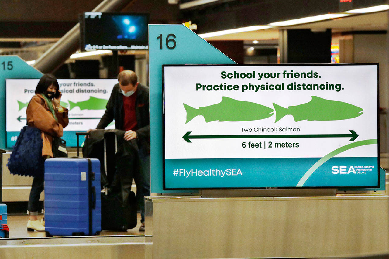 Travelers pick up luggage where signs remind them, with classic Pacific Northwest icons showing the size of two Chinook salmon, to stay six feet apart at Seattle-Tacoma International Airport on Monday, May 18, 2020, in SeaTac. Monday was the first day that travelers at the airport were required to wear face coverings in the public areas there. The Port of Seattle has encouraged its employees to wear face coverings, and all federal agencies that operate at the airport require their employees to wear them. All airlines operating at SeaTac require employees and passengers to wear face coverings. (Elaine Thompson/Associated Press)