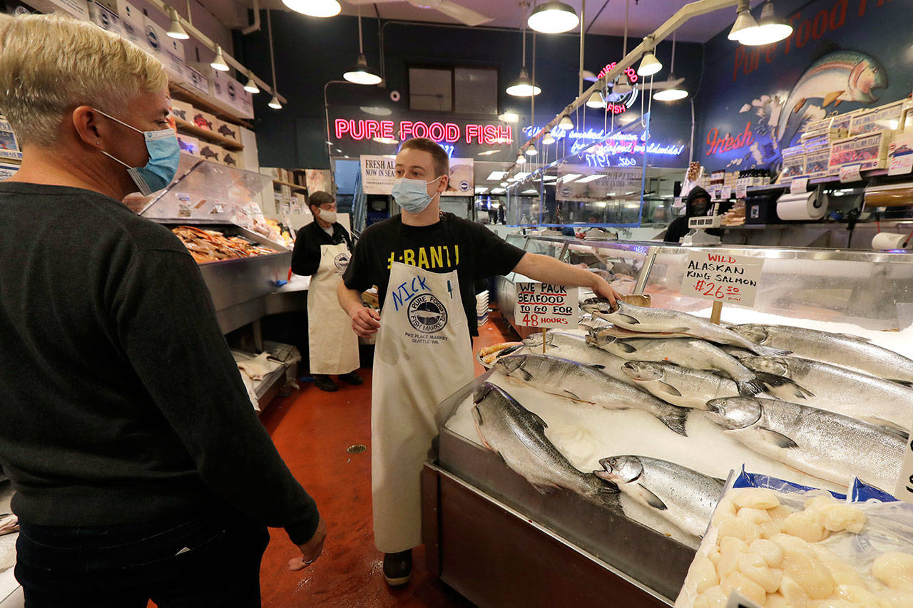 Nicholas Hartmann, center, shows a fish to a customer as he works at the Pure Food Fish Market on Tuesday, July 7, 2020, at Pike Place Market in Seattle. Tuesday was the first day of a new statewide order that requires people to wear masks or other facial coverings inside businesses in hopes of slowing the spread of the coronavirus. Business owners who fail to refuse service to customers who don’t wear masks can face fines or lose their business license, but some business owners have raised concerns about turning away customers. (Ted S. Warren/Associated Press)