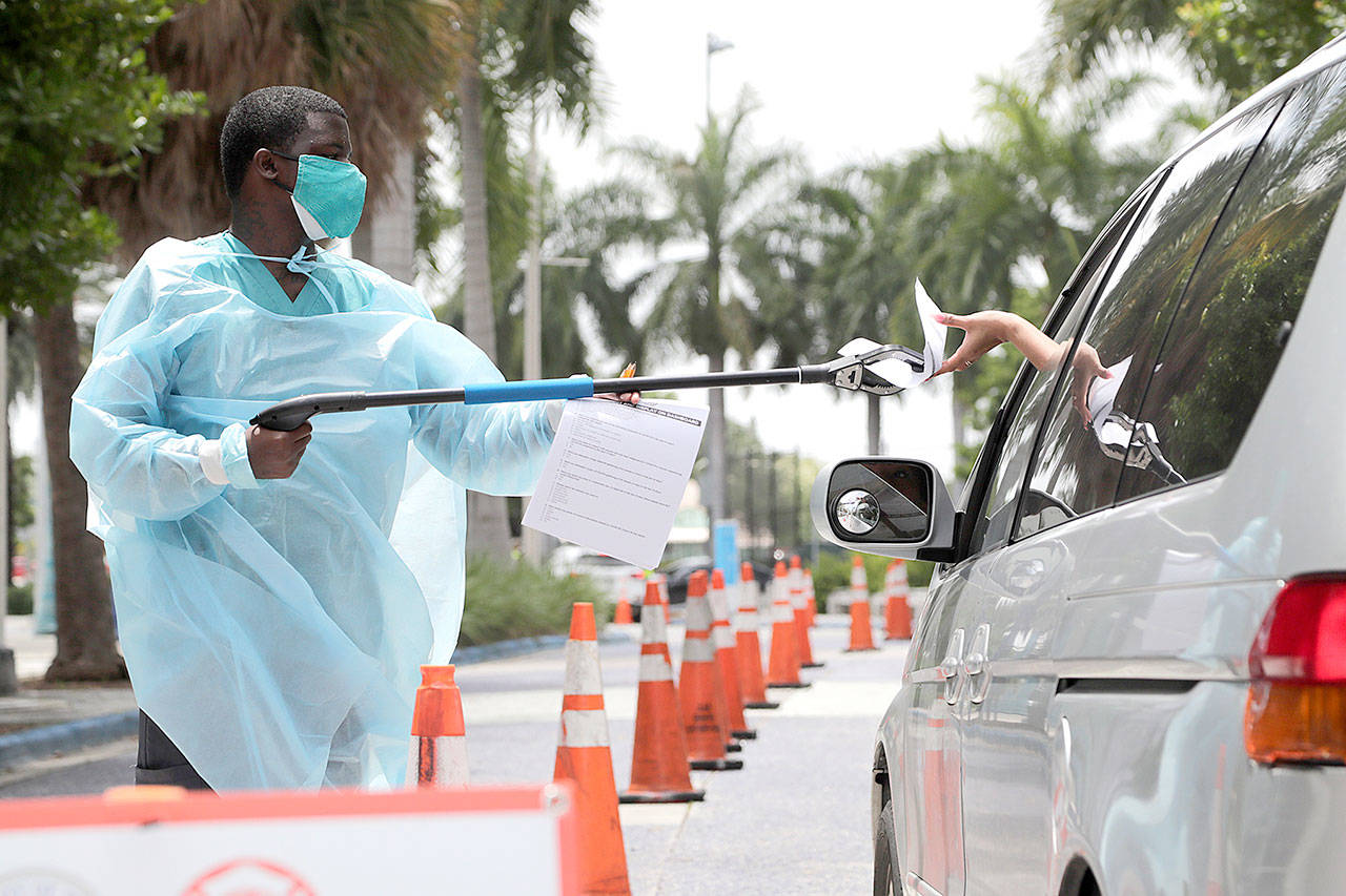 Health care worker Dante Hills, left, passes paperwork to a woman in a vehicle at a COVID-19 testing site outside of Marlins Park on Monday, July 27, 2020, in Miami. The Marlins home opener against the Baltimore Orioles on Monday night has been postponed as the Marlins deal with a coronavirus outbreak that stranded them in Philadelphia. (Lynne Sladky/Associated Press)