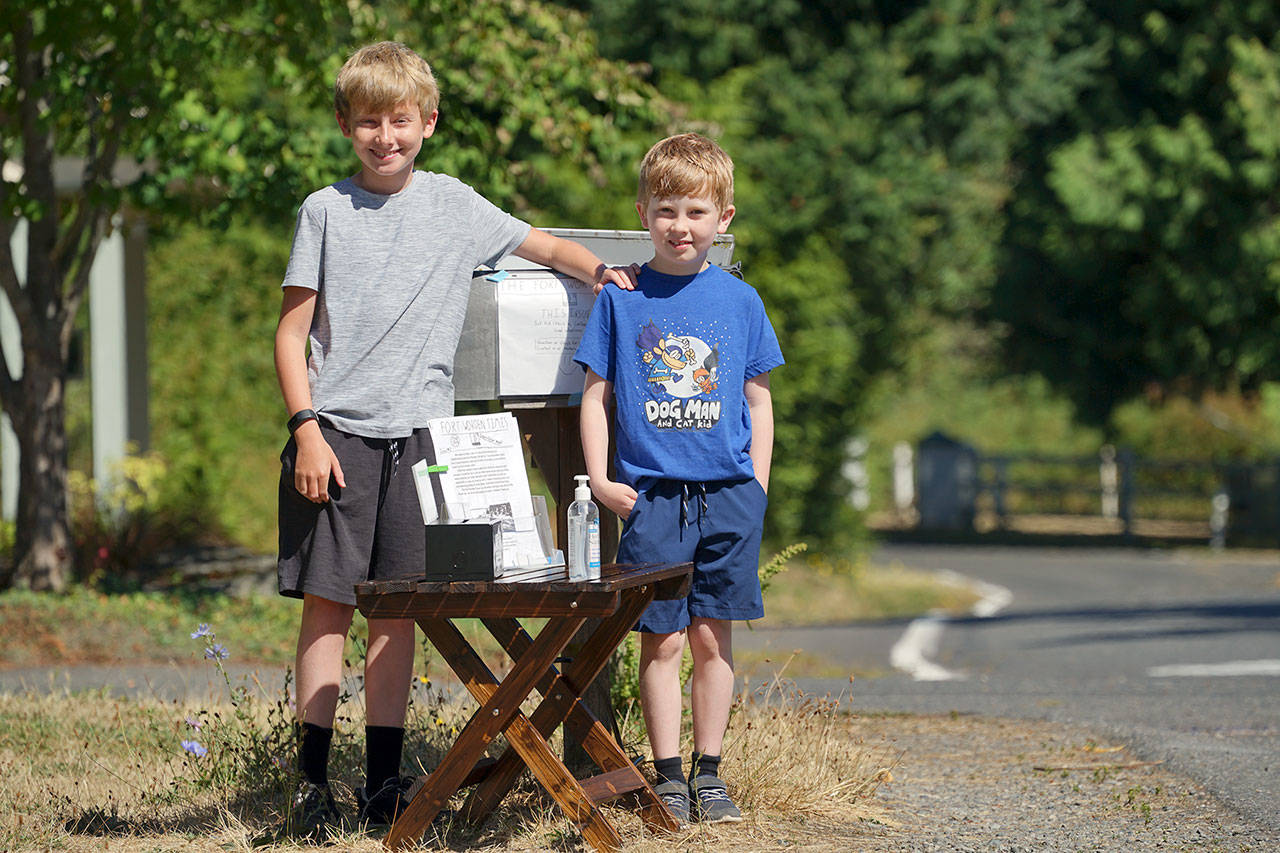 Noah Isenberg, 11, left, and Elliot Isenberg, 7, stand next to their newspaper stand at the corner of Spruce and W streets in Port Townsend. Growing restless while stuck at home during the pandemic, the brothers decided to create The Fort Worden Times. (Nicholas Johnson/Peninsula Daily News)