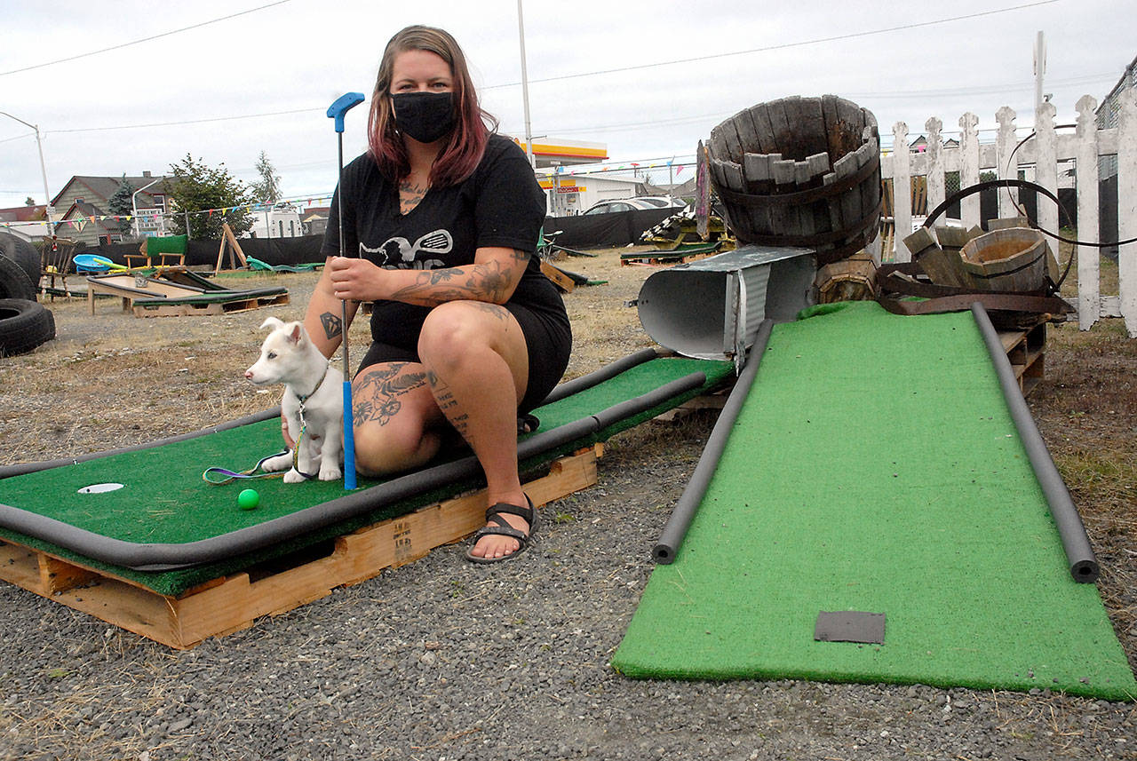 OMG Olympic Miniature Golf owner Molly Barnes, along with her dog, Elsa, kneels at the first hole of a course built primarily of scavanged materials on Tuesday in Port Angeles. (Keith Thorpe/Peninsula Daily News)