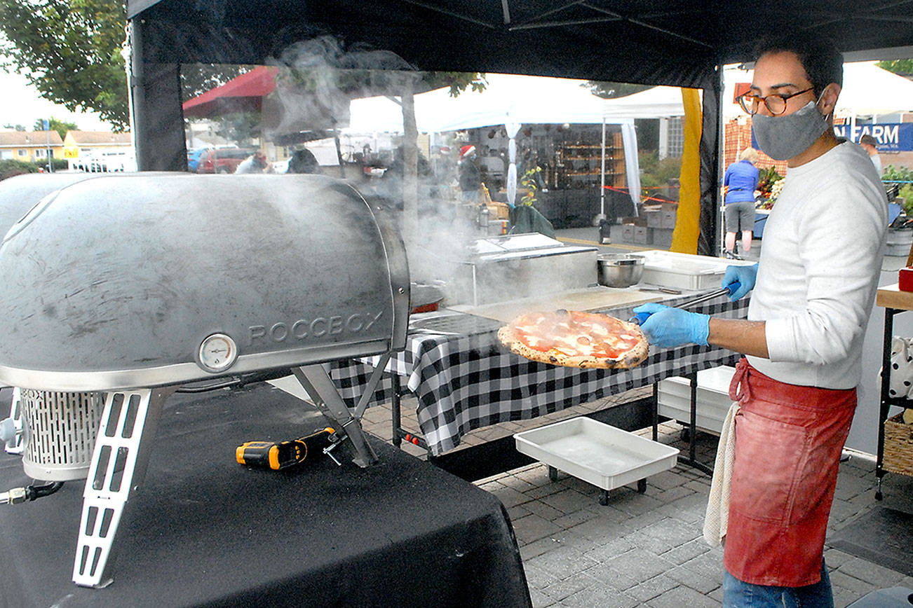 Keith Thorpe/Peninsula Daily News Hot wares Andréa Mingiano of Sequim-based Ulivo Pizzeria pulls a steaming hot pizza from a gas fired oven at his vendor’s tent at last Saturday’s Sequim Farmers and Artisans Market. The market operates from 9 a.m. to 3 p.m. Saturdays on the plaza of the Sequim Civic Center at Cedar Street and North Sequim Avenue. This Saturday, the Sequim Irrigation Festival will host the Innovative Arts and Crafts Fairat the market. For more about the festival, which is largley online this year, see irrigationfestival.com.