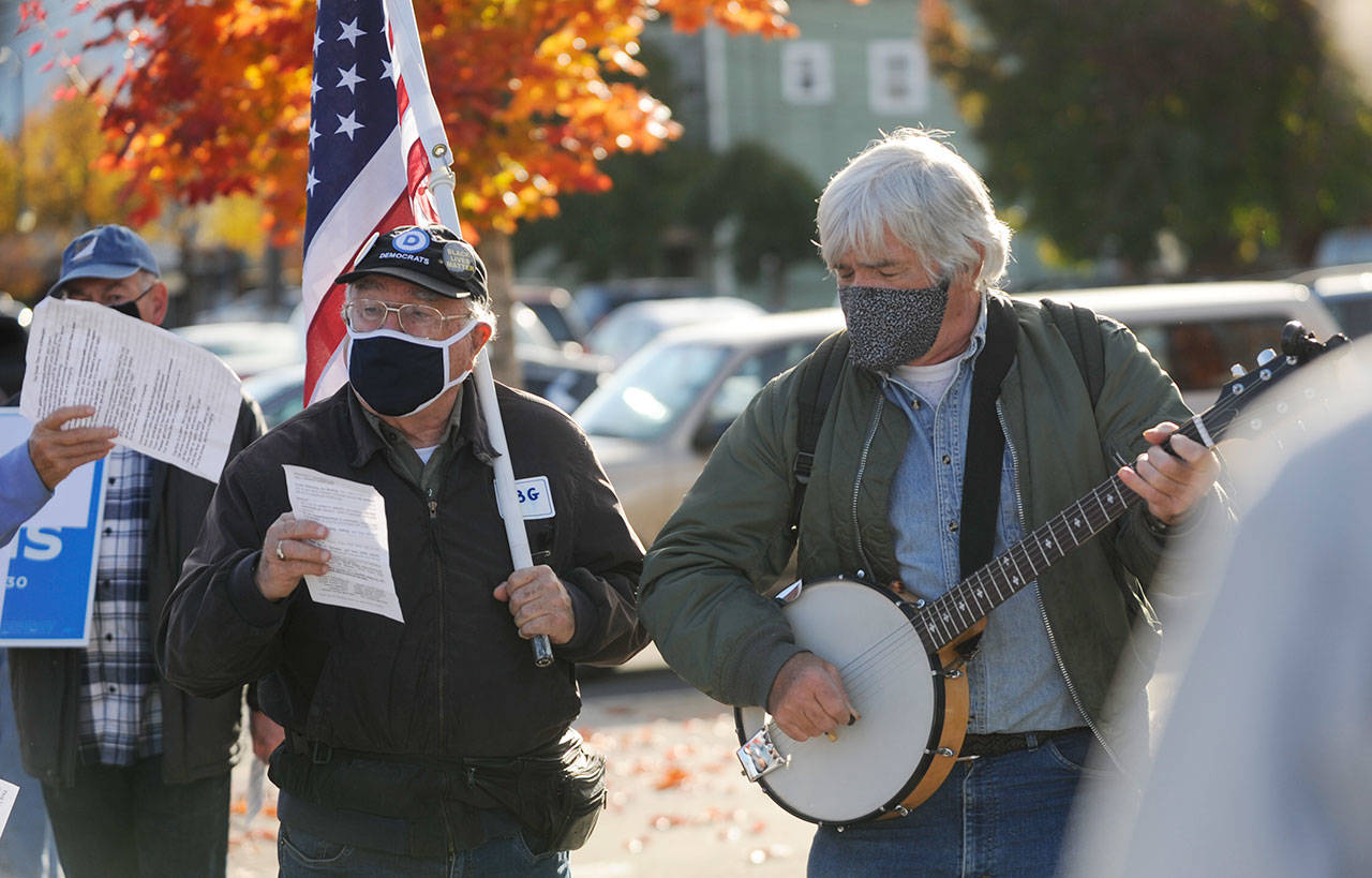 Tim Wheeler, left, and a group of about 40 individuals sing Woody Guthrie’s “This Land Is Your Land,” accompanied by Steve Koehler on banjo, at the Sequim Civic Center on Wednesday afternoon. The pair joined an event organized in part by Indivisible Sequim and part of a nation-wide rally to protect the results of the 2020 general election in case President Donald Trump refuses to concede. (Michael Dashiell/Olympic Peninsula News Group)