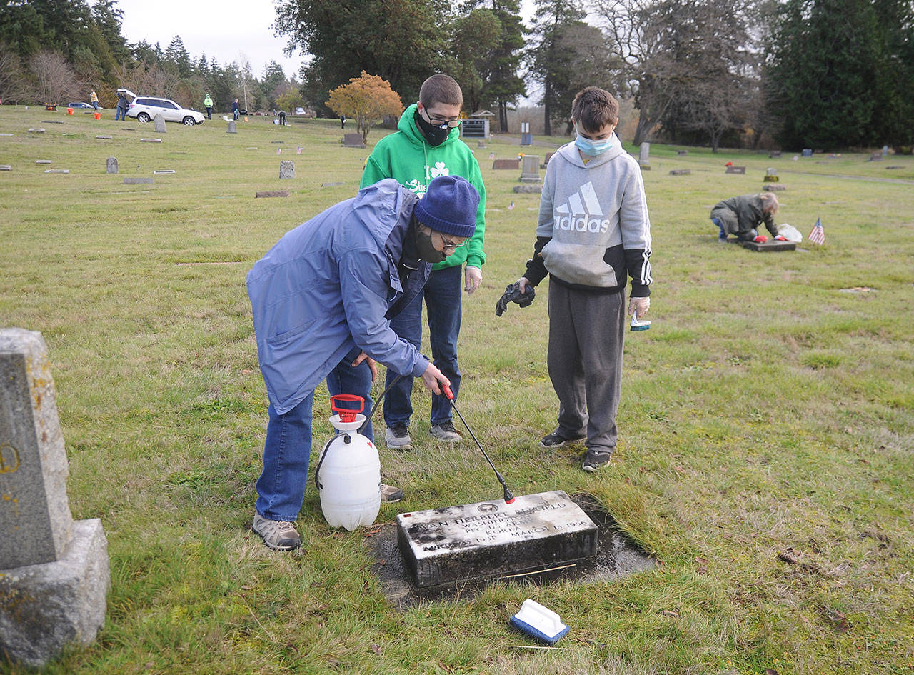 Jan Urfer, secretary with the Michael Trebert Chapter of the Daughter of the American Revolution, works with scouts Hunter Halverson and Cayden Beauregard from Sequim Troop 90 to clean headstones and grave markers at Sequim View Cemetery on Nov 28. Members of the chapter and scout troop were busy preparing the cemetery for a Wreaths Across America event slated for Dec. 19. (Michael Dashiell/Olympic Peninsula News Group)