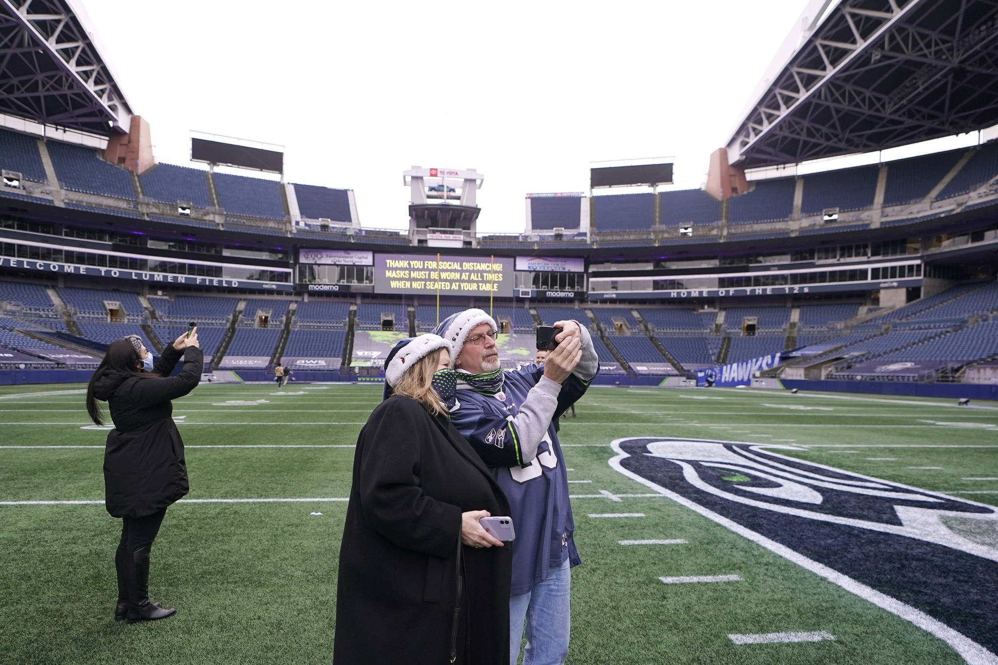 Tom Gallagher, right, wears a Seattle Seahawks jersey and holiday hat as he takes a photo with his wife Debbie on the turf at Lumen Field, Thursday, Feb. 18, 2021, in Seattle. The couple, who are Seattle Seahawks season ticket holders, were celebrating their 45th anniversary by taking part in the "Field To Table" event at the Seahawks' home stadium on the first night of several weeks of dates that offer four-course meals cooked by local chefs and served at socially distanced tables in an open-sided tent on the field as a precaution against the COVID-19 pandemic. (AP Photo/Ted S. Warren)