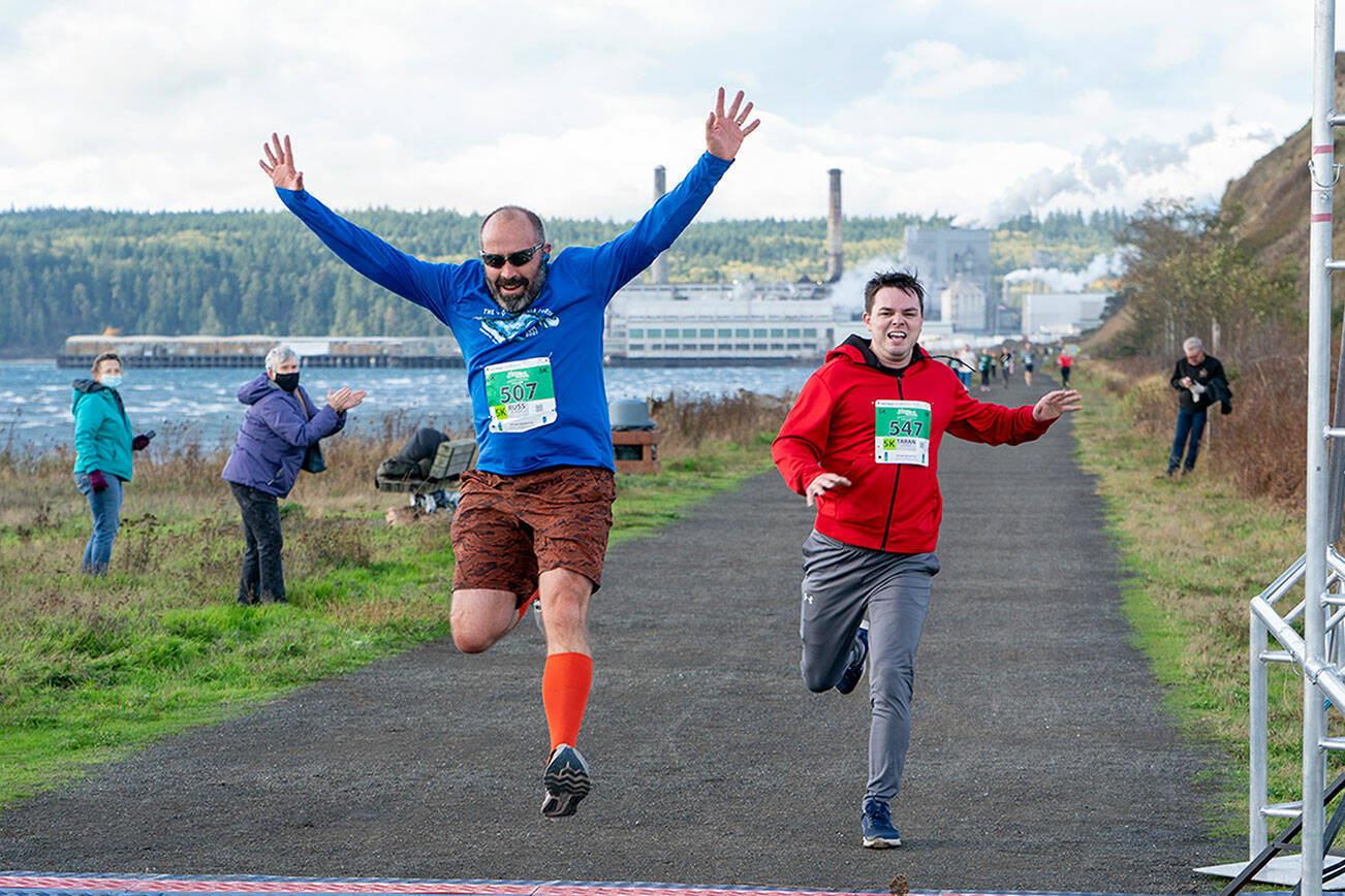 Sequim’s Russ Britton (507) jumps through the finish line after he completes the Run the Peninsula 5K on the Larry Scott Trail in Port Townsend on Saturday. Taran Johnson, (547) also from Sequim, races to the finish. (Steve Mullensky/for Peninsula Daily News)