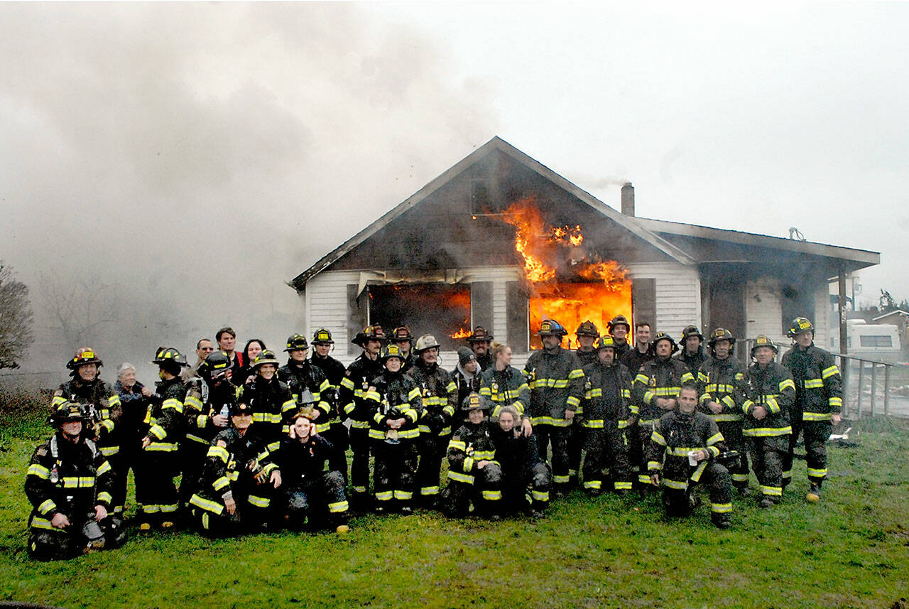 Firefighters and medics from Clallam County Fire District 2 pose for photos in front of a flaming house, part of a training exercise on Sunday at 2354 E. Fifth Ave. in the Gales Addition east of Port Angeles. The burn of the donated house allowed firefighters to hone their skills in a live-fire scenario through two days of training sessions. Once extinguished, the site will be cleared to make way for future development. (Keith Thorpe/Peninsula Daily News)
