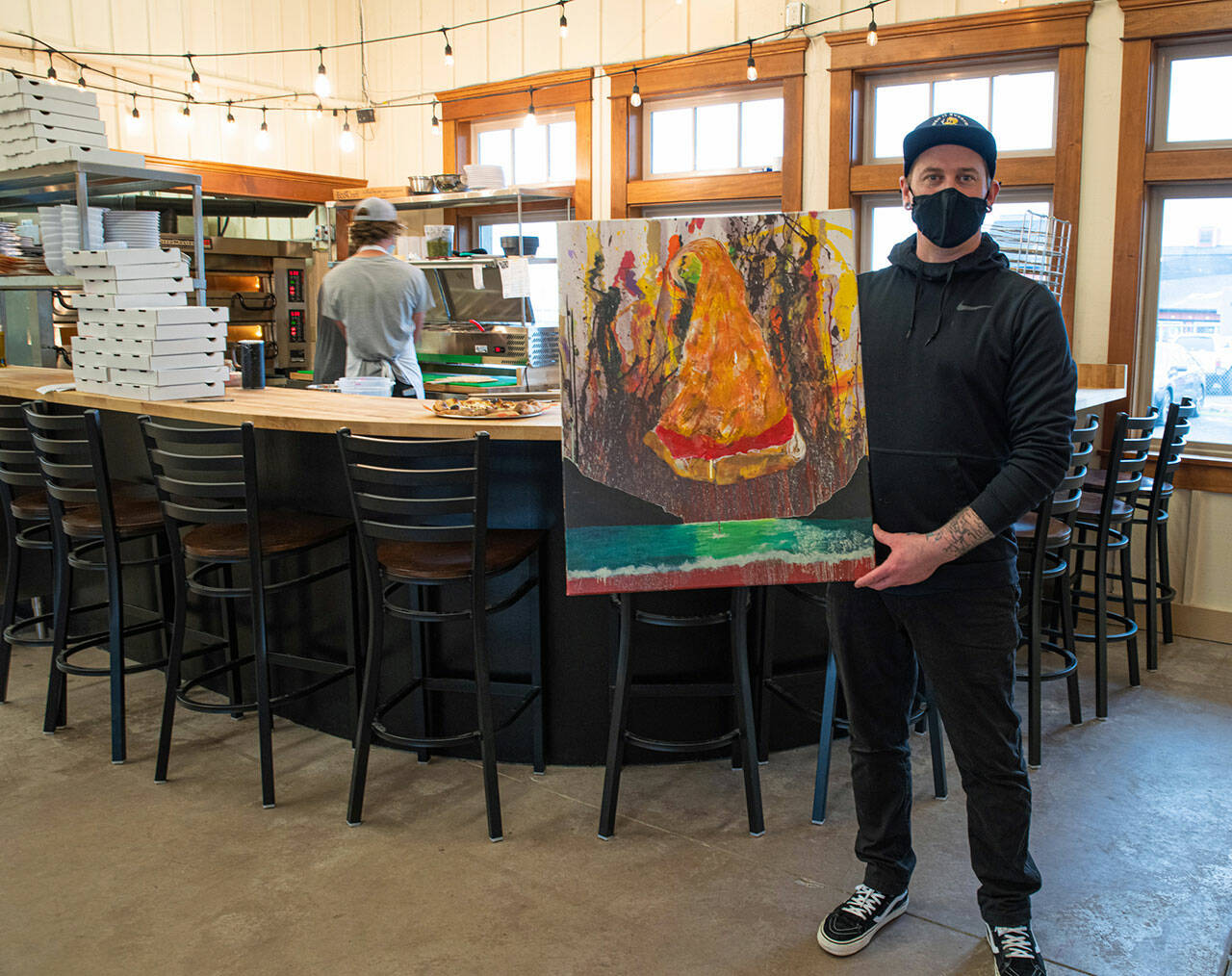 Larry White, district manager for Barhop Brewing & Artisan Pizza and artist, holds a painting for Barhop's new Sequim location.Behind him, kitchen manager Jeffrey Adams prepares pizzas for customers in the open kitchen. (Emily Matthiessen/Olympic Peninsula News Group)