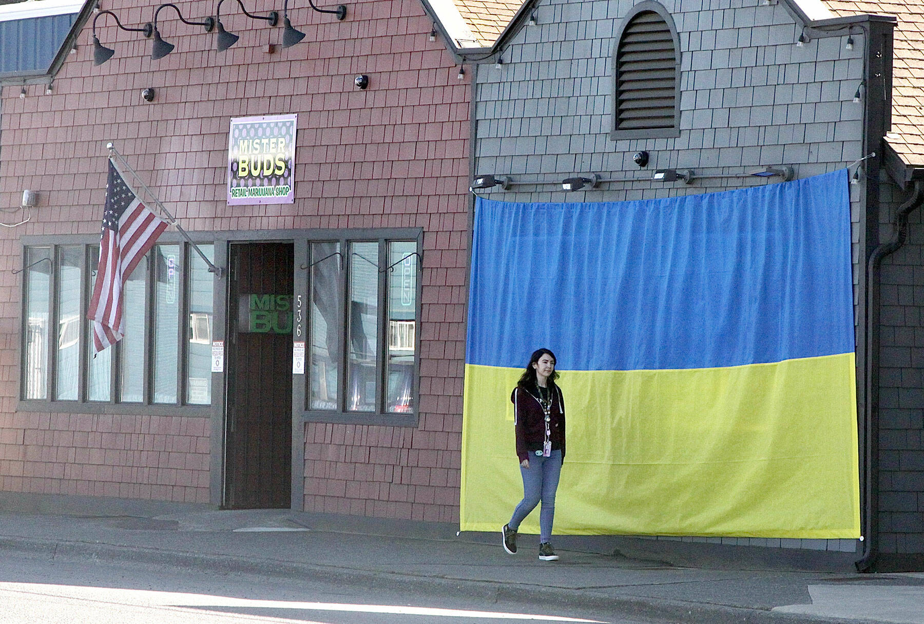 Shawna Small walks on Marine Drive in Port Angeles past a large Ukrainian flag on the outside of Mister Buds, which owner Malik Atwater put up recently along with the U.S. flag he has flown for many years. The flag was hand-sewn by Vivian Wai, Atwaters wife, after the two discussed how to support the Ukrainians as they fight against the invasion of the country launched by Russia on Feb. 24. We felt helpless to do anything to help, Atwater said, adding that it was Wai who came up with the idea. (Dave Logan/for Peninsula Daily News)