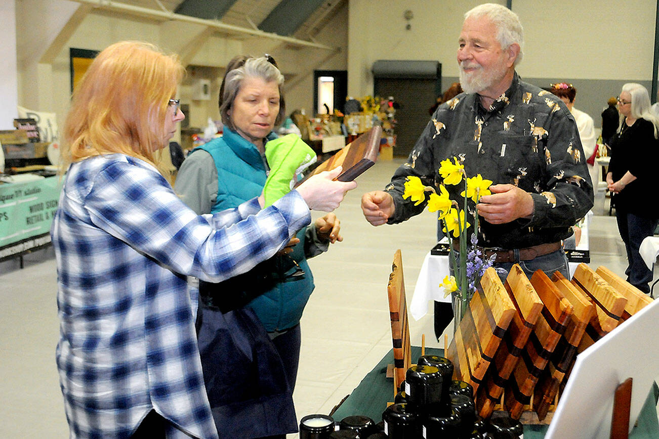 Keith Thorpe/Peninsula Daily News
Cari Gavin, left, and Pam Wennerberg, both of Port Angeles, examine a cutting board created by Tim Smith of Port Angeles-based Blacktail Trail LLC,  right, during Saturday's Mother's Day Market at Vern Burton Community Center in Port Angeles. The event featured a variety of vendor displays with items created mostly by local artisans.