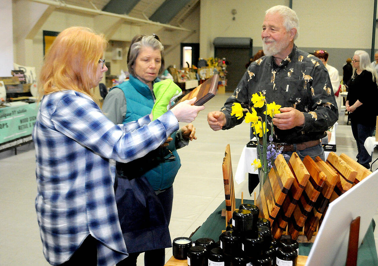 Cari Gavin, left, and Pam Wennerberg, both of Port Angeles, examine a cutting board created by Tim Smith of Port Angeles-based Blacktail Trail LLC, right, during Saturday’s Mother’s Day Market at Vern Burton Community Center in Port Angeles. (Keith Thorpe/Peninsula Daily News)