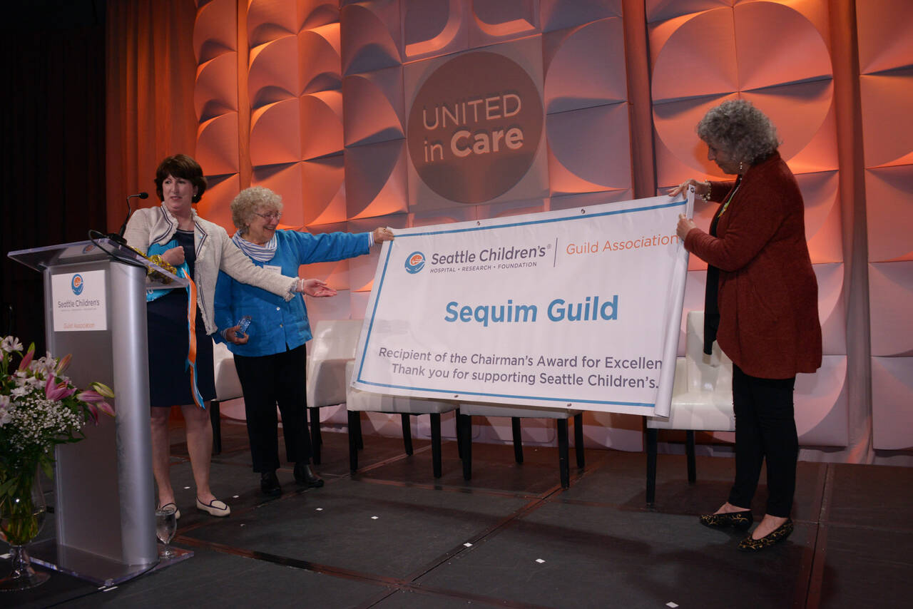 Ann Shanahan, a trustee with the Seattle Children’s Guild Association, presents a banner displaying the Chairman’s Award to Sequim Guild President Kathy Bare and Secretary Lydia Stanhope at the Seattle Children’s Guild Association 2022 Annual Meeting & Luncheon on May 13. (Courtesy photo)