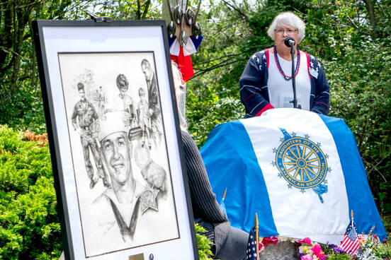 Joan Shields Bennett, wife of former Navy Seabee Marvin Shields, reads her remarks to assembled guests during a re-dedication of a plaque honoring her late husband, who died in Vietnam in 1965. Shields, portrayed in the drawing on the stand, was awarded the Congressional Medal of Honor for actions above and beyond the call of duty. The original plaque, made of bronze, was stolen in 2019 and was never recovered. (Steve Mullensky/for Peninsula Daily News)