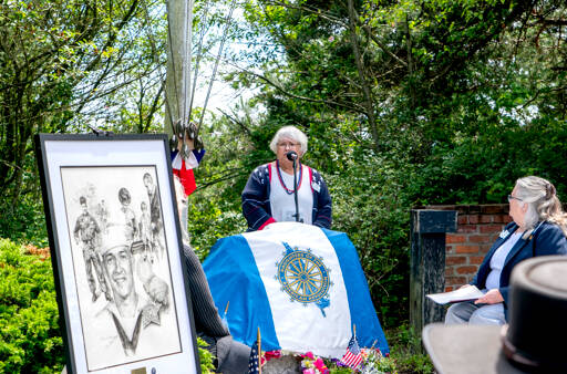 Joan Shields Bennett, wife of former Navy Seabee Marvin Shields, reads her remarks to assembled guests during a re-dedication of a plaque honoring her late husband, who died in Vietnam in 1965. Shields, portrayed in the drawing on the stand, was awarded the Congressional Medal of Honor for actions above and beyond the call of duty. The original plaque, made of bronze, was stolen in 2019 and was never recovered. (Steve Mullensky/for Peninsula Daily News)