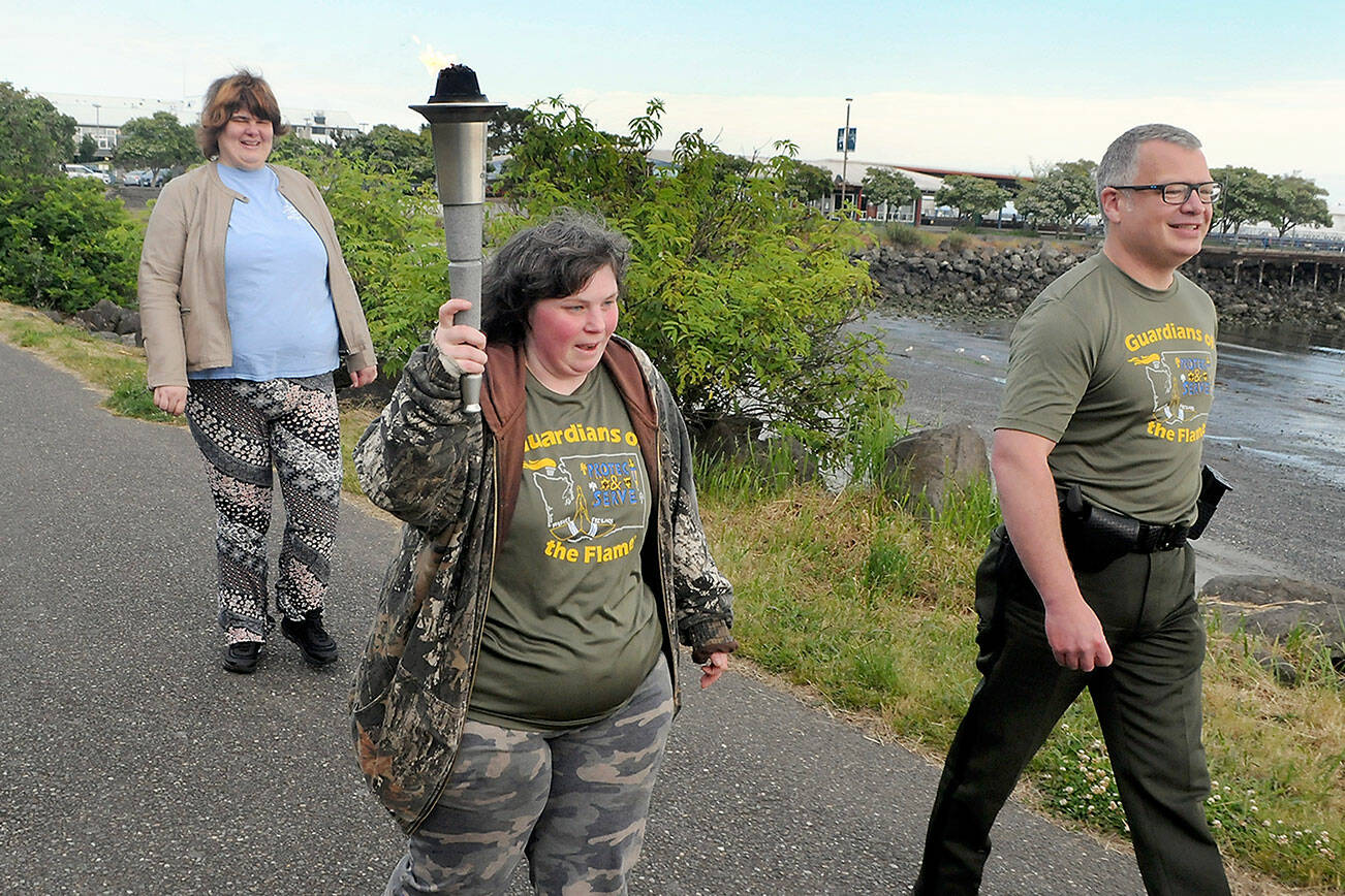 Special Olympian Deedra Hunter of Port Angeles takes a turn carrying the torch accompanied by fellow Olympian Bonny Ann Cates, left, and Chief Criminal Deputy Brian King of the Clallam County Sheriff’s Office during Wednesday’s Law Enforcement Torch Run along a section of the Waterfront Trail in Port Angeles. The relay, which included representatives from the sheriff’s offices in Clallam and Jefferson counties, Port Angeles and Sequim police, tribal police from Lower Elwha Klallam and Jamestown S’Klallam, State Patrol, Quilcene firefighters, U.S. Border Patrol, state parks personnel and Olympic National Park rangers, as well as several Special Olympians, followed a route from the west side of Port Angeles at 7 a.m. to the Hood Canal Bridge to about 5 p.m. in support of Special Olympics, which provides sports training and athletic competition for individuals with disabilities. (Keith Thorpe/Peninsula Daily News)