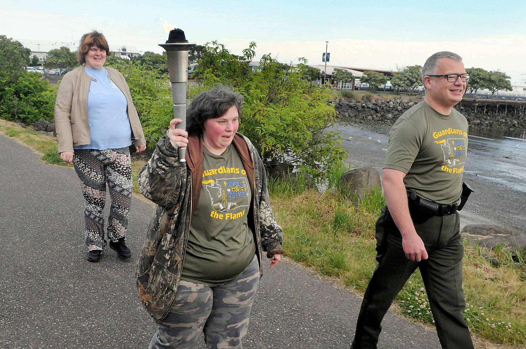 Special Olympian Deedra Hunter of Port Angeles takes a turn carrying the torch accompanied by fellow Olympian Bonny Ann Cates, left, and Chief Criminal Deputy Brian King of the Clallam County Sheriffs Office during Wednesdays Law Enforcement Torch Run along a section of the Waterfront Trail in Port Angeles. The relay, which included representatives from the sheriffs offices in Clallam and Jefferson counties, Port Angeles and Sequim police, tribal police from Lower Elwha Klallam and Jamestown SKlallam, State Patrol, Quilcene firefighters, U.S. Border Patrol, state parks personnel and Olympic National Park rangers, as well as several Special Olympians, followed a route from the west side of Port Angeles at 7 a.m. to the Hood Canal Bridge to about 5 p.m. in support of Special Olympics, which provides sports training and athletic competition for individuals with disabilities. (Keith Thorpe/Peninsula Daily News)