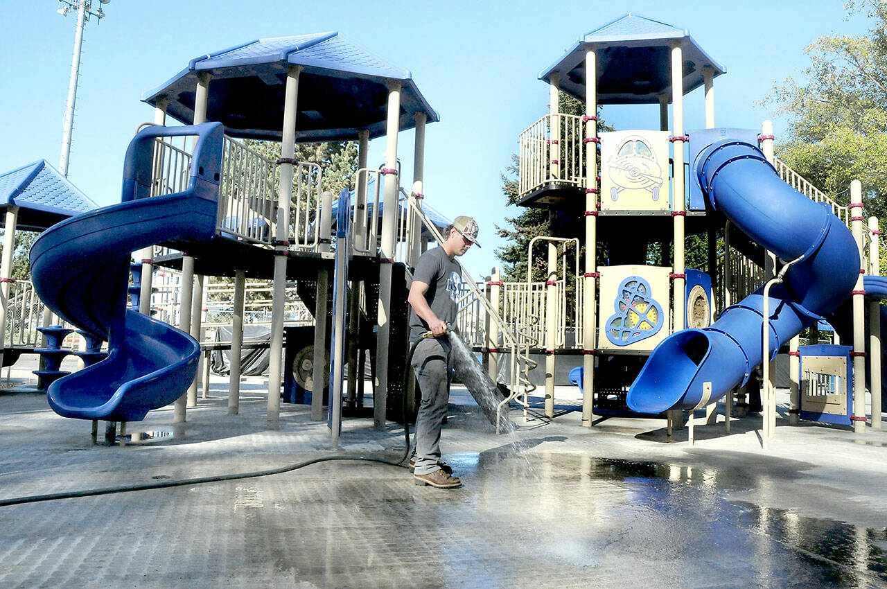 Port Angels Parks & Recreation Department employee Easton Goslin hoses down the concrete underlayment at the playground at Shane Park on the west side of Port Angeles on Wednesday. The playground was closed in mid-September after tiles of rubberized material that made up the play surface began to come loose, creating a hazard to play equipment users. The old tiles have been removed and are awaiting replacement. The playground area remains closed until work can be completed. (Keith Thorpe/Peninsula Daily News)