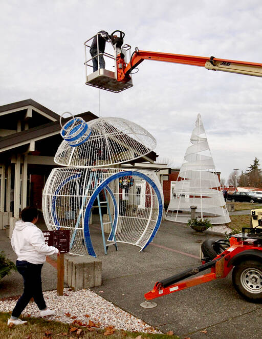 An oversized ornament for the opening ceremony at 5 p.m. Wednesday for the 32nd annual Festival of Trees this coming weekend is put in place on Sunday at the Vern Burton Community Center in Port Angeles, with volunteer Ricki Smith in the bucket, Matt Williams working inside the 14-foot-tall ornament and Laci Williams watching. Four of the huge round ornaments, a 20-foot-tall tree and two fountains donated by Microsoft to the Olympic Medical Center Foundation, will be lit up outside the community center during a free 20-minute ceremony that also will include performances by the Port Angeles Symphony, Ballet Workshop and Ghostlight Productions. A limited number of tickets, which will provide seats under cover outside and a sneak preview of the trees to be auctioned off on Friday, are available for $20 by going to omcf.org. (Dave Logan/for Peninsula Daily News)