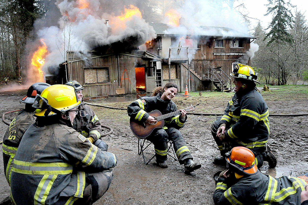 Clallam County Fire District 2 firefighter Mya Delano plays her guitar surrounded by fellow squad members as a house at 141 Harry Brown Road burns behind her on Sunday west of Port Angeles. The house was intentionally torched as a training exercise, giving firefighters an opportunity to practice in a live fire situation. Fire districts 2 and 4, along with members of the Port Angeles Fire Department, took part in the event. (Keith Thorpe/Peninsula Daily News)