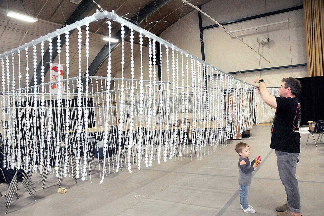 Ryan Schroeder of Port Angeles makes adjustments to a beaded hanging ceiling decoration as his son, Noah, 3, looks on before the hanging was hoisted into place during Thursday’s preparation for the 30th annual Juan de Fuca Festival of the Arts at Vern Burton Community Center in Port Angeles. The three-day festival kicks off today at Vern Burton and Port Angeles City Hall, and it spreads out to additional venues on Saturday and Sunday. (Keith Thorpe/Peninsula Daily News)