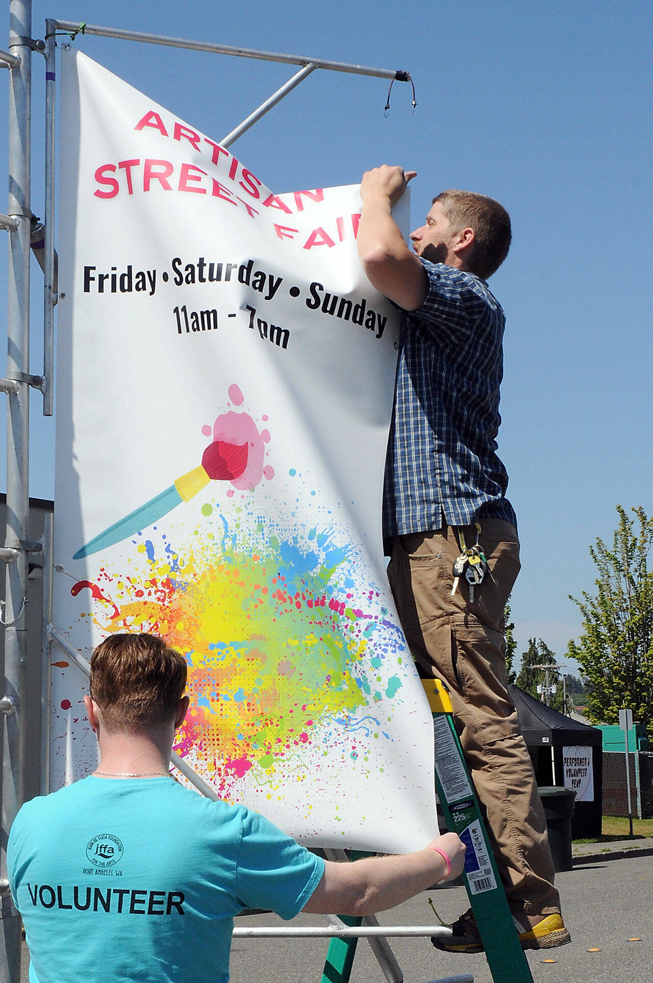 Silas Crews of Sequim, right, hoists a Juan de Fuca Festival Artisan Street Fair banner with the assistance of Morgan Martholick of Port Angeles on Friday outside of Vern Burton Community Center in Port Angeles. (KEITH THORPE/PENINSULA DAILY NEWS)