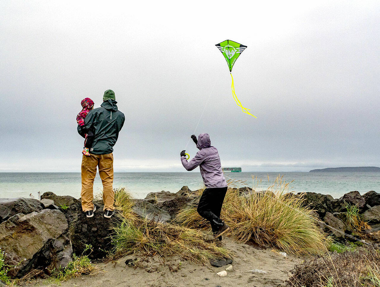 Mara, 3, held by her father Mike Karas, watch as her mother, Molly, launches a kite into the 15 mph winds off the rocks at the Point Wilson Lighthouse on Sunday at Fort Worden State Park. (Steve Mullensky/for Peninsula Daily News)