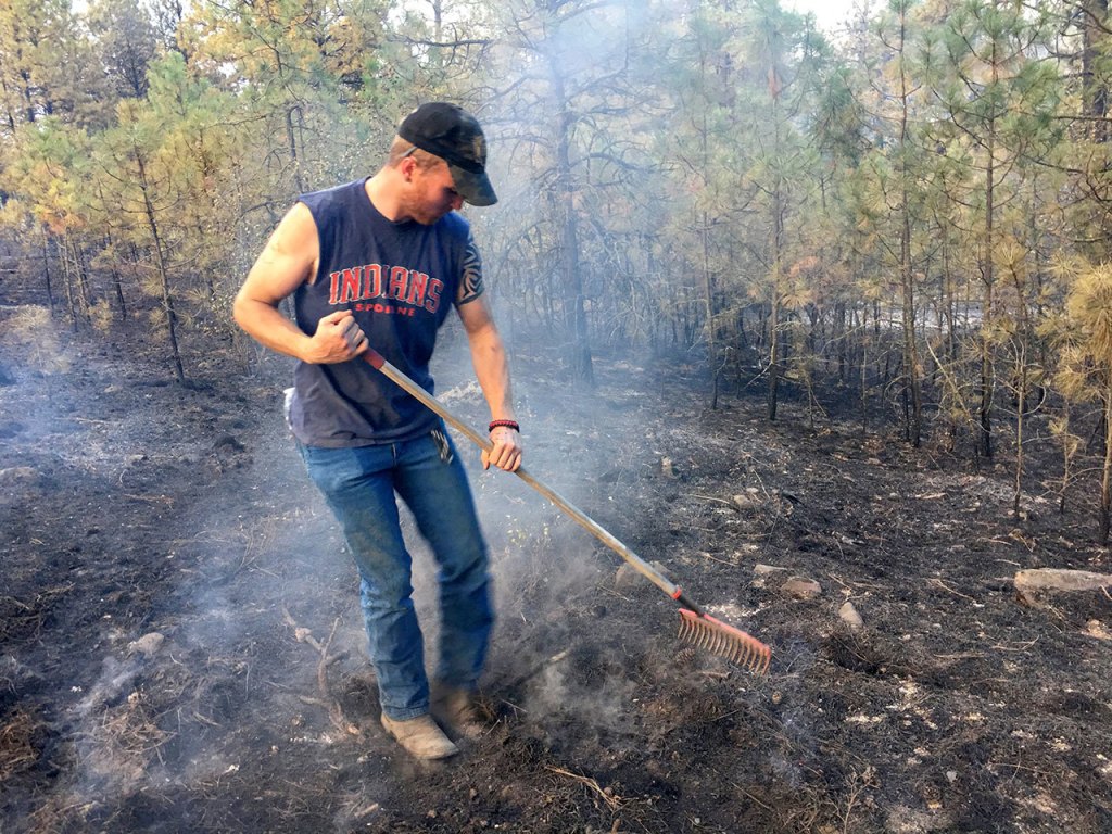 In the aftermath of a fire that started from a downed power line, JJ Anderson uses a rake to put out hot spots on the property of his brother PJ Anderson, located northeast of Spangle on Sunday. His brother&rsquo;s home was spared, but others were not so lucky. (Jesse Tinsley/The Spokesman-Review, via AP)