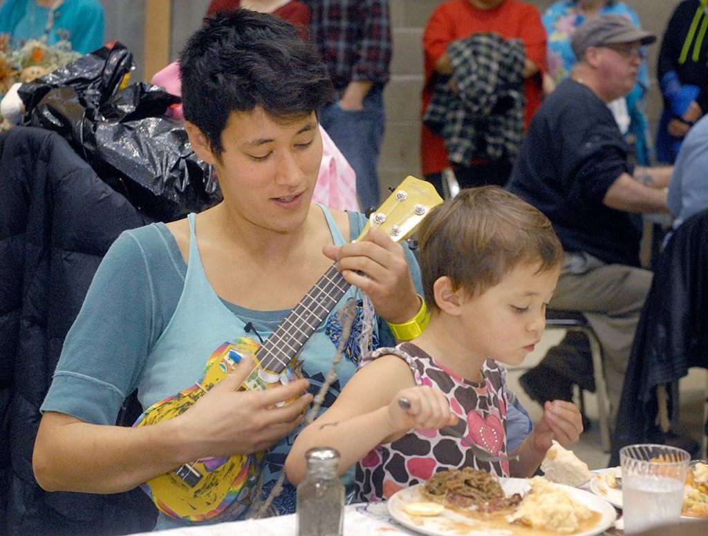 Taz Love of Port Angeles plays the ukulele as her daughter, Shalom Love, 3, eats dinner at Thursday&rsquo;s community Thanksgiving meal at Queen of Angels Catholic Church in Port Angeles. (Keith Thorpe/Peninsula Daily News)