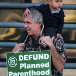 Ed Carver, holding his son, protests outside a Planned Parenthood health center in the Van Nuys section of Los Angeles on Saturday. Several dozen protesters gathered in California&rsquo;s San Fernando Valley demanding the organization be stripped of its federal funding. (Richard Vogel/The Associated Press)