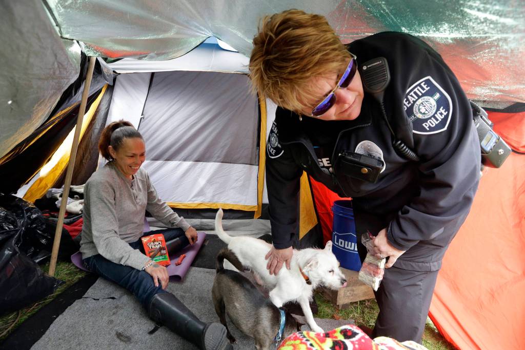 In this photo taken March 23, Seattle Police Officer Tawnia Pfaff, right, hands out biscuits to dogs belonging to Sheryl, a homeless woman who declined to give her last name, as part of a new team of outreach workers and officers that go out and connect homeless people to services in Seattle. Sheryl has lived in a tent in a greenbelt near Interstate Highway 90 for about six months. (Elaine Thompson/The Associated Press)