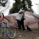 In this photo taken March 23, Lisa Hooper rakes debris from in front of the tent where she lives in a greenbelt near a freeway, as another camper wheels a bicycle past her in Seattle. (Elaine Thompson/The Associated Press)