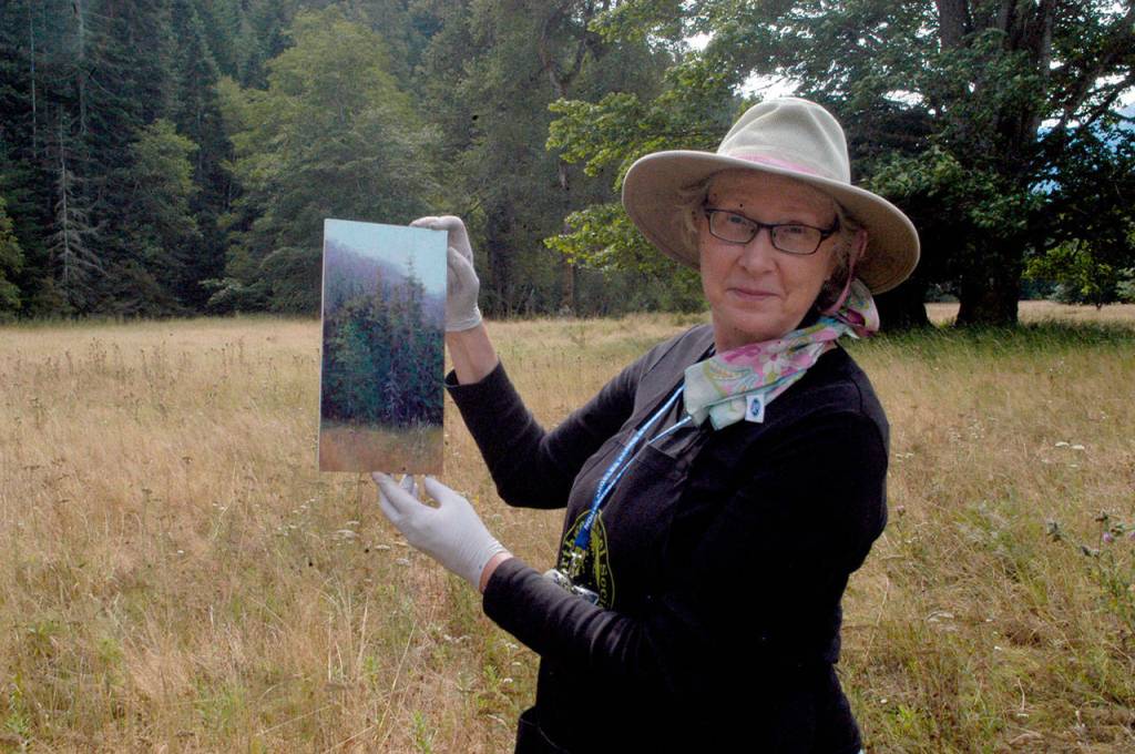 Gretha Lindwood poses with her completed plein art landscape painting in a meadow across from the Elwha River while participating in Paint the Peninsula on Wednesday. (Sarah Sharp/Peninsula Daily News)