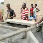 ABOVE: Justin Adjogble, far left, supervises a crew as they work on a Dignity Toilet in Zogbedgi. Along with family members who are being given the toilet can be seen Taouvik Boukari, third from left, and Kolawole Dokou, far right.                                RIGHT: The main residential road in Zogbedgi is packed dirt. Drains from outdoor shower stalls keep the roadway wet even in the dry season.