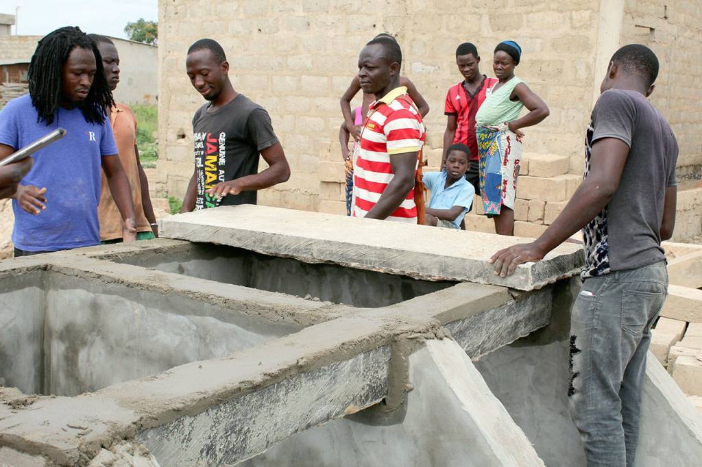 ABOVE: Justin Adjogble, far left, supervises a crew as they work on a Dignity Toilet in Zogbedgi. Along with family members who are being given the toilet can be seen Taouvik Boukari, third from left, and Kolawole Dokou, far right.                                RIGHT: The main residential road in Zogbedgi is packed dirt. Drains from outdoor shower stalls keep the roadway wet even in the dry season.