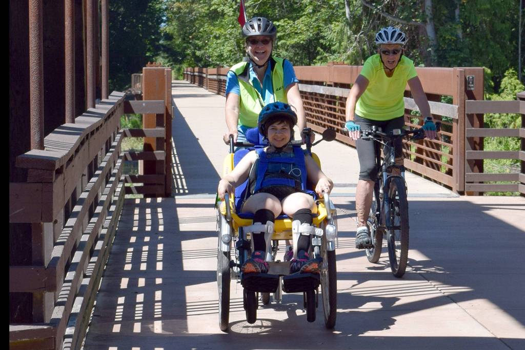 Sequim Wheelers volunteer Elaine Cates pilots a wheelchair bike with Clallam Mosaic participant Kaitlyn Winn, foreground, and Sequim Wheelers volunteer and safety Michele Fraker, right, at Railroad Bridge Park.