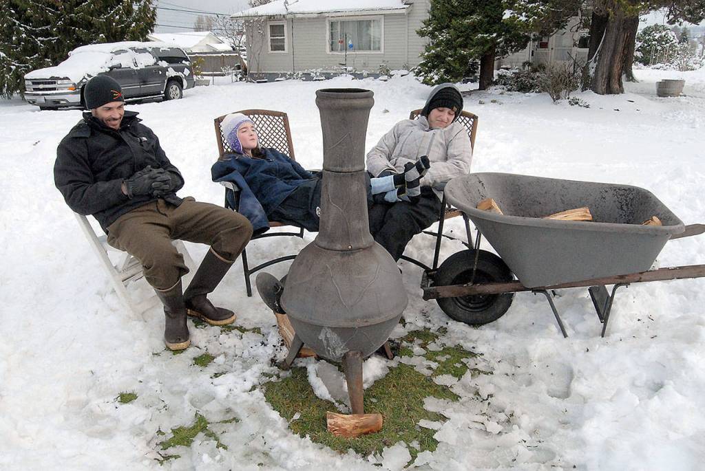 Not detoured by a landscape covered with a blanket of snow, Dan Callahan, Brenna Callahan, 10, and Emily Callahan bask in the heat of a portable wood stove set up next to the street near their home at Fifth and Oak streets in Port Angeles on Saturday. Dan Callahan said it was an ideal way to spend an afternoon people watching and enjoying the cold winter air. (Keith Thorpe/Peninsula Daily News)