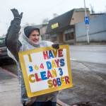 Jenni Tiderman stands in the pouring rain at the corner of First and Lincoln Streets in Port Angeles on Friday with a sign declaring she is 364 days sober. Over the last year she has secured housing and a job. She also is regaining custody of her children. (Jesse Major/Peninsula Daily News)
