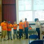 Anastasia Rigby announces information about an item being introduced for auction. To her left are four Spartan senior boys who volunteered 22 hours of work together to be auctioned off. On the screen in the background is a projection of bids in process. (Zorina Barker/for Peninsula Daily News)